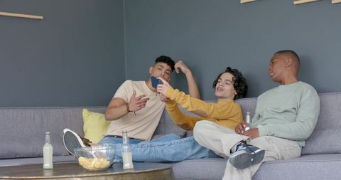 Multicultural men relaxing on sofa sharing smartphone and snacks in casual home hangout
