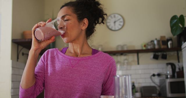 Woman drinking pink strawberry smoothie in home kitchen, promoting healthy eating and wellness. Ideal for content related to healthy habits, nutrition, lifestyle blogs, and kitchen settings.