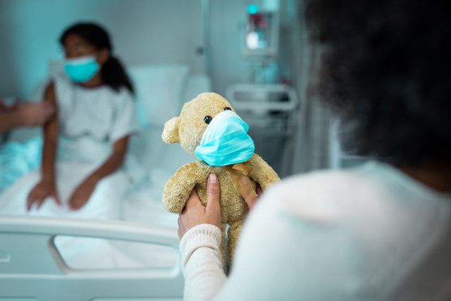 This image shows an African American mother and daughter sitting on a hospital bed, both wearing face masks. The daughter is holding a teddy bear also wearing a face mask. This image can be used in articles or advertisements related to healthcare, pediatric care, family support during illness, COVID-19 safety measures, and hospital environments.