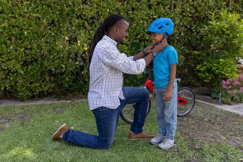 African american father fastening bright blue helmet on son in backyard before bike ride