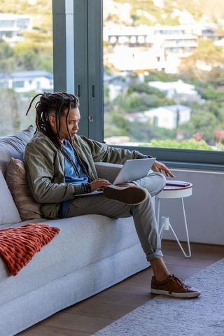 African american man with dreadlocks sitting on sofa typing on laptop overlooking hillside