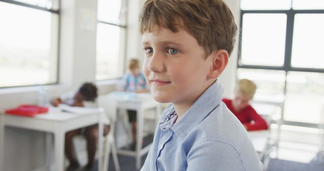 Young boy sitting in a classroom, looking thoughtful, with classmates studying in the background. Ideal for educational materials, school-related promotions, and children's development articles.