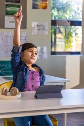 Young student raising hand in modern classroom using tablet and headphones, engaged learning