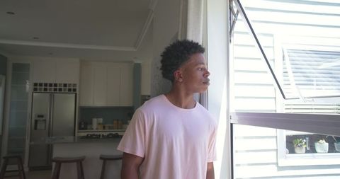 Young man standing by open window in modern kitchen, looking outside, contemplative mood