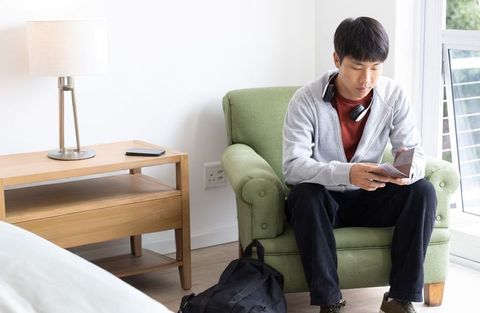 Asian man sitting on green armchair holding smartphone and wallet in bright bedroom