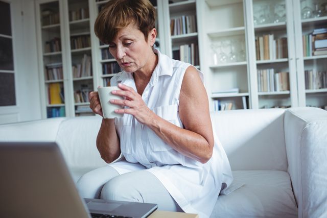 Senior woman enjoying a cup of coffee while using a laptop in a cozy living room. Ideal for themes related to senior lifestyle, technology use among elderly, home comfort, and casual relaxation. Suitable for articles, blogs, and advertisements focusing on senior citizens, home living, and technology adoption.