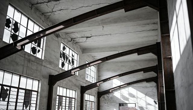 Arched steel beams showing rust and broken windows in abandoned factory hall with sunlight
