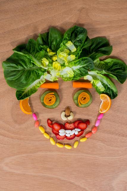 This image shows a smiley face made from various vegetables and confectionery items arranged on a wooden table. The face includes leafy greens for hair, carrot slices for eyes, kiwi slices for pupils, an almond for a nose, strawberry slices and beans for a mouth, and colorful candies for decoration. Ideal for use in articles or advertisements promoting healthy eating, creative food presentation, children's nutrition, or fun food ideas.