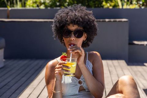 African american woman sipping citrus cocktail on rooftop lounge wearing sunglasses