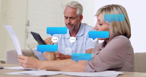 Senior couple reviewing bills and reading messages on tablet while sorting paperwork at home