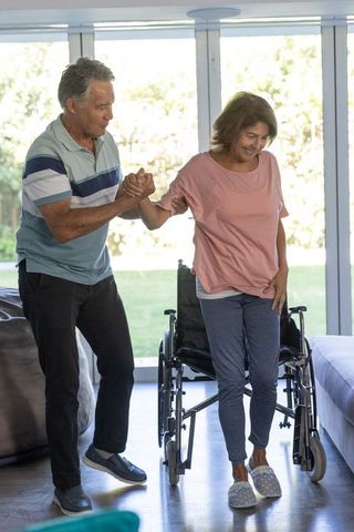 Mature man assisting senior woman standing from wheelchair in bright living room