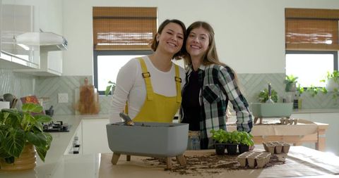 Smiling diverse women potting basil in sunny kitchen, indoor gardening and plant care