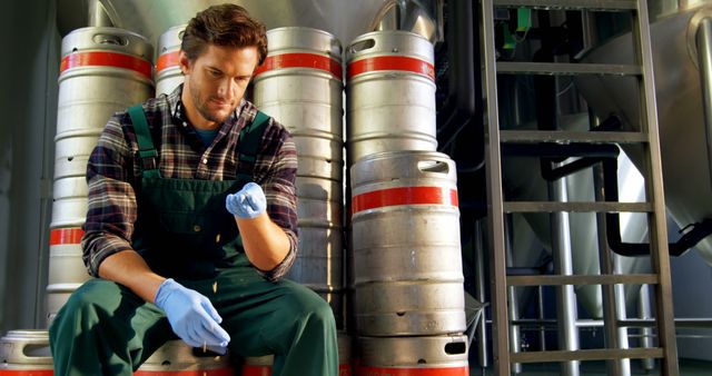 Brewery worker in gloves checking quality control of beer process in warehouse. Surrounded by metal barrels. Useful for industrial, brewing, and beverage production industry content. Ideal for articles on quality assurance, brewery tours, and labor in beer manufacturing.