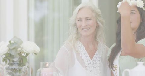 Senior woman smiling at bridal shower while friend adjusting floral crown and sash, soft pastel rose