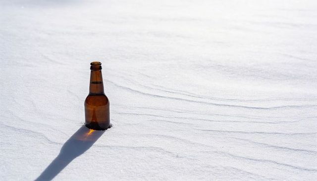 Amber bottle sinking into sunlit rippled snow casting long shadow minimal winter solitude