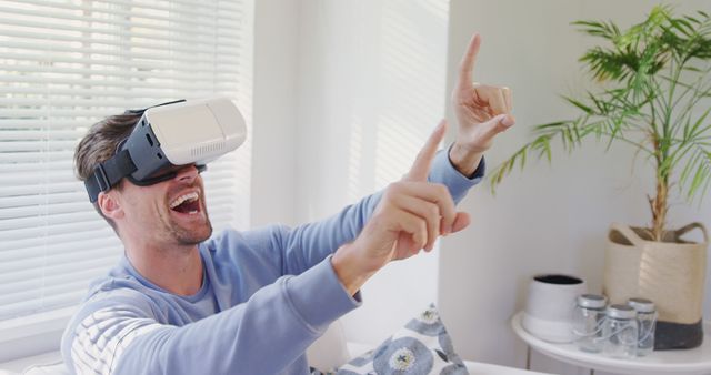 Young man sitting on a couch at home experiencing virtual reality, laughing and pointing while wearing a VR headset. Perfect for advertising VR technology, gaming experiences, or showcasing the fun and engaging aspects of immersive tech. Suitable for tech blogs, VR product promotions, or lifestyle articles about virtual reality.