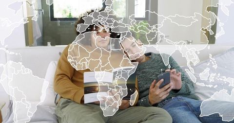 Two men relaxing on couch reading booklet, scrolling smartphone with world map overlay