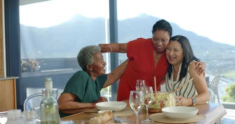 Multigenerational women embracing and laughing around dining table sharing salad and wine