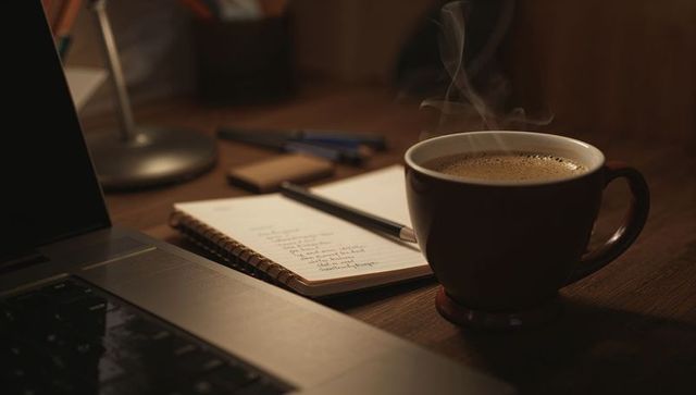 Steaming coffee cup on wooden desk with notebook pen and laptop for cozy evening workspace