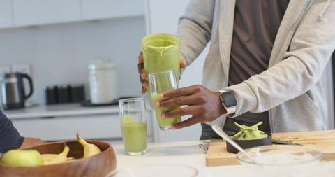 Pouring green smoothie into tall glasses on kitchen island, friends making healthy drink