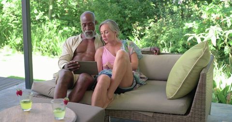 Mature multiracial couple relaxing on covered patio sofa sharing tablet and drinks