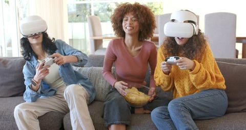 Diverse friends playing multiplayer VR games on couch sharing snacks and laughing together
