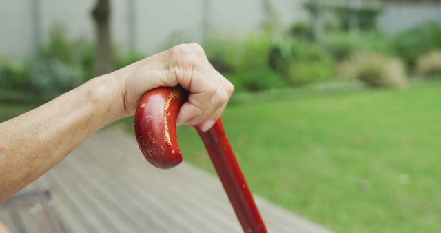 Shows an elderly hand holding a red walking cane in an outdoor setting. Ideal for use in articles or promotions related to senior care, mobility aids, elderly independence, or retirement living.