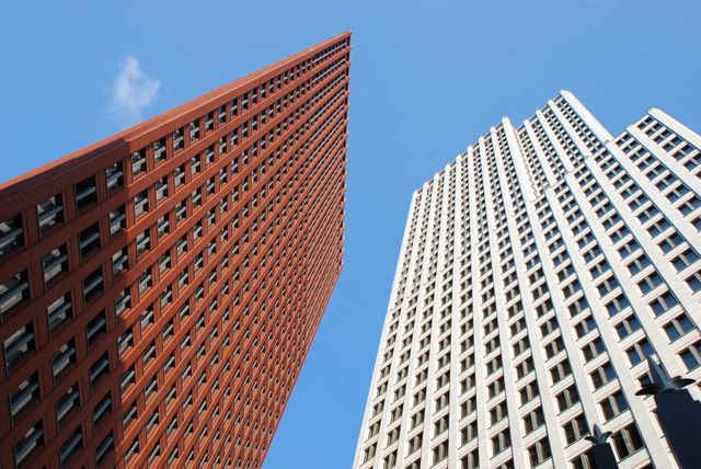 This image showcases two high-rise office buildings photographed from below, emphasizing their geometric patterns and height against a clear blue sky. The contrasting colors and shapes of the red and beige buildings create a striking visual composition ideal for use in urban planning, architectural design presentations, real estate marketing, or city-themed artwork.