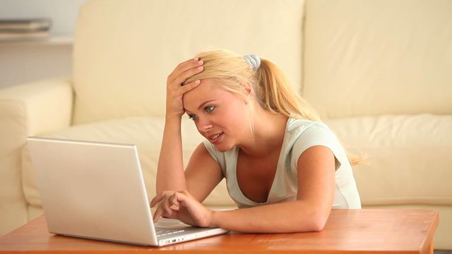 Blonde woman sitting on wooden table in the living room, using a laptop and smiling while chatting. Perfect for illustrating concepts such as online communication, remote friendship, modern technology use, and home lifestyle.