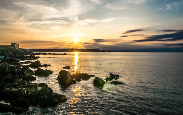 Sunset reflecting off calm sea with rocky shoreline, creating peaceful scene. Ideal for travel websites, blogs, calendars, motivational posters focusing on nature or tranquility.