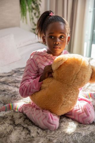 Contemplative african american girl hugging teddy bear in sunlit pink pajamas