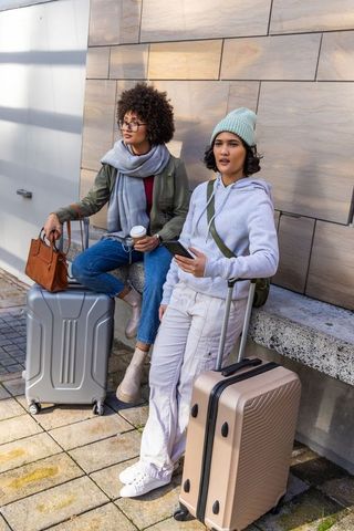 Diverse female travelers waiting with rolling suitcases, coffee and smartphone in urban plaza