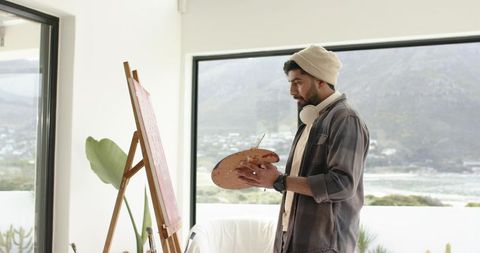 Indian artist painting on canvas at easel with coastal view, wearing beanie and headphones