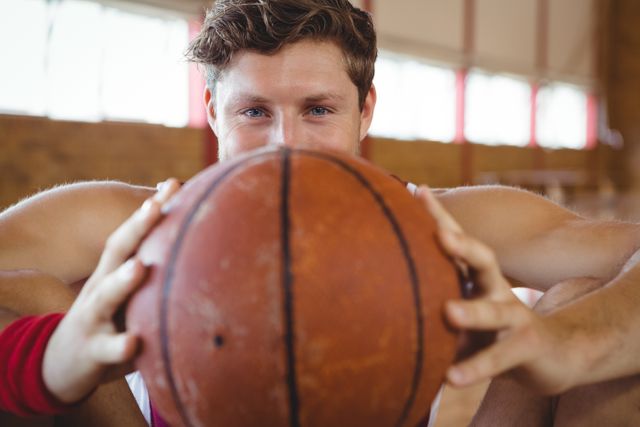 Young male basketball player sitting on court, holding ball close to face, showing determination and focus. Ideal for sports promotions, fitness campaigns, athletic training programs, and motivational posters.