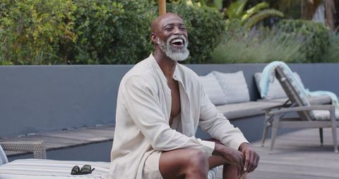 Laughing senior black man relaxing poolside on deck wearing light linen shirt, summer leisure