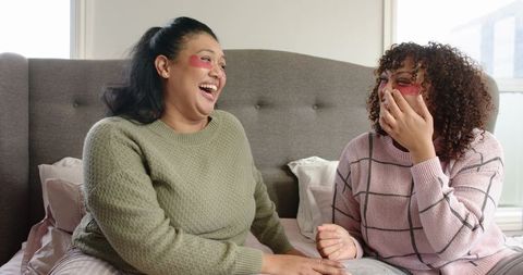 Two friends laughing during cozy self-care night with red under-eye gel patches on bed