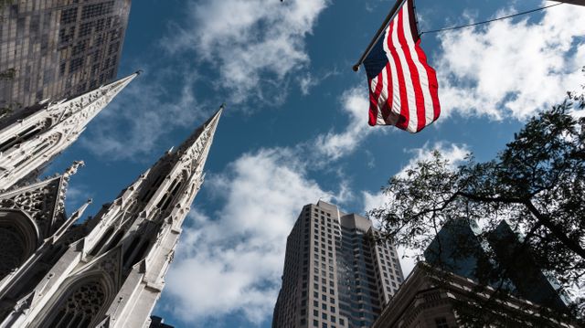This image features an American flag prominently displayed with tall skyscrapers against a blue sky with clouds. Suitable for themes related to patriotism, urban life, architecture, and American cities. Can be used for articles, blog posts, or websites related to American culture, travel, tourism, or national holidays like Independence Day and Memorial Day.