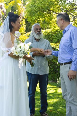 Bride exchanging rings with asian groom during outdoor garden wedding officiant passing ring