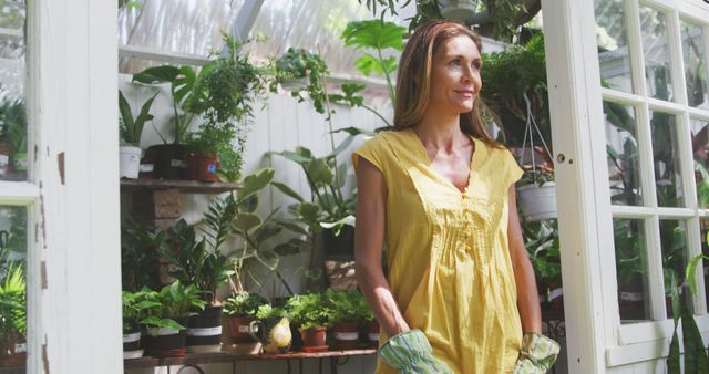 Happy caucasian woman wearing gloves standing in greenhouse in sunny garden. Nature, gardening and hobbies.