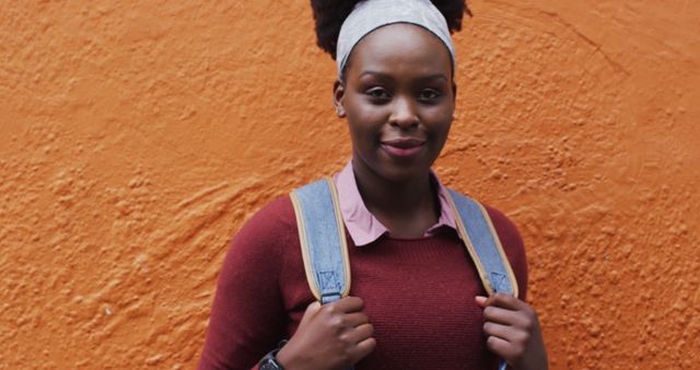 Young woman with afro hair and headband stands confidently against a bright orange textured wall, smiling while holding the straps of her denim backpack. Perfect for themes related to youth, urban lifestyle, positivity, and personal confidence. Can be used in blogs, advertisements, social media posts, and educational content.