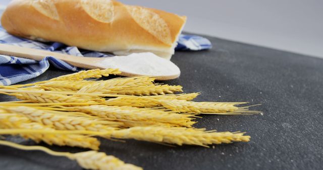 Freshly baked baguette lying on blue and white cloth, accompanied by wooden spoon filled with flour and golden wheat ears on black surface. Ideal for use in articles, blogs, and advertisements related to baking, food industry, French cuisine, and farm-to-table ingredients.