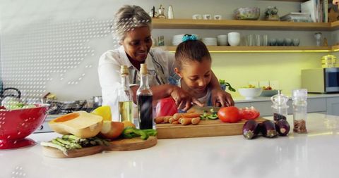 Grandmother guiding granddaughter chopping vegetables on kitchen island