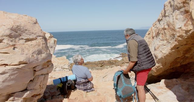 Back view of diverse senior couple talking on rocks by sea. Retirement, vacations, togetherness, summer, active senior lifestyle, unaltered.