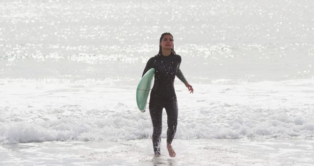 Surfer confidently walking out of ocean waves, holding surfboard, wearing a wetsuit. Ideal for themes related to surfing, adventure sports, and coastal travel. Suitable for use in marketing campaigns for surfing gear, travel blogs, and lifestyle magazines focused on outdoor activities.