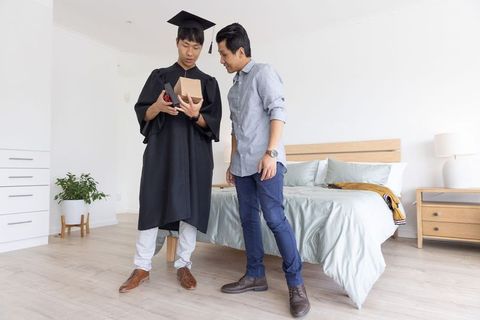 Asian father and graduate son celebrating home graduation opening gift and checking phone