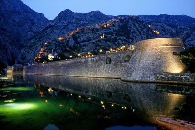 Spectacular night view of Kotor Fortress reflected in calm water, illuminated against Montenegro's mountain backdrop. Ideal for travel brochures, historical documentaries, tourism adverts, and articles on medieval architecture.