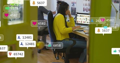 Young woman working at laptop in open plan office with social media notification overlays