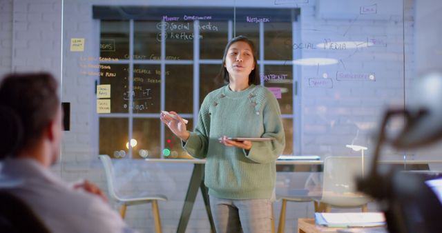 Businesswoman giving a presentation to her team in a modern office with a glass wall covered in notes and ideas. She holds a tablet while explaining social media strategies. Ideal for content about professional development, creative workplaces, teamwork, marketing strategies, and business planning.