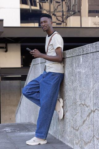 Urban man leaning on concrete wall checking smartphone in striped tee and wide denim jeans