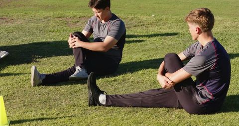 Diverse male teammates stretching on grass hugging knees during outdoor team training